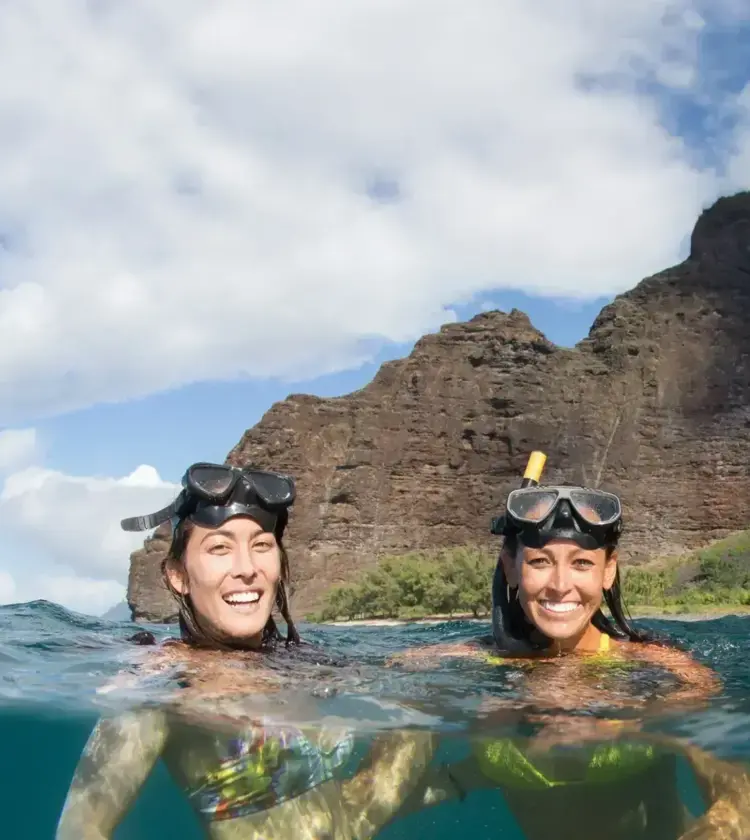 two women snorkeling in the ocean near volcanic cliffs under partly cloudy sky
