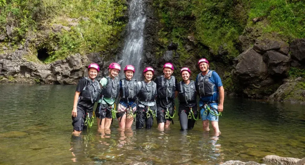 Seven adventure tourists in helmets standing in shallow water by a waterfall in a rocky, green landscape