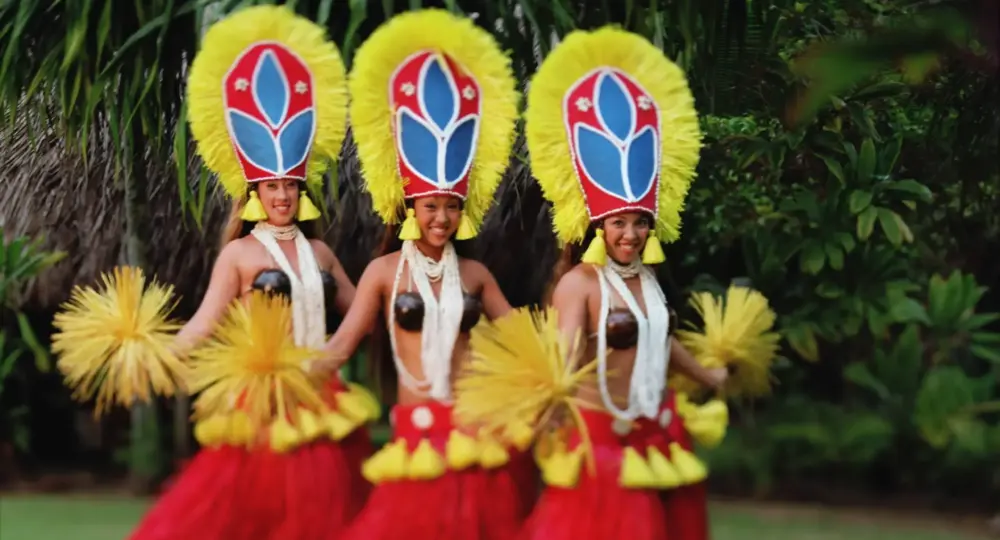 Three Hawaiian hula dancers with yellow feathered headdresses and red grass skirts dancing outdoors