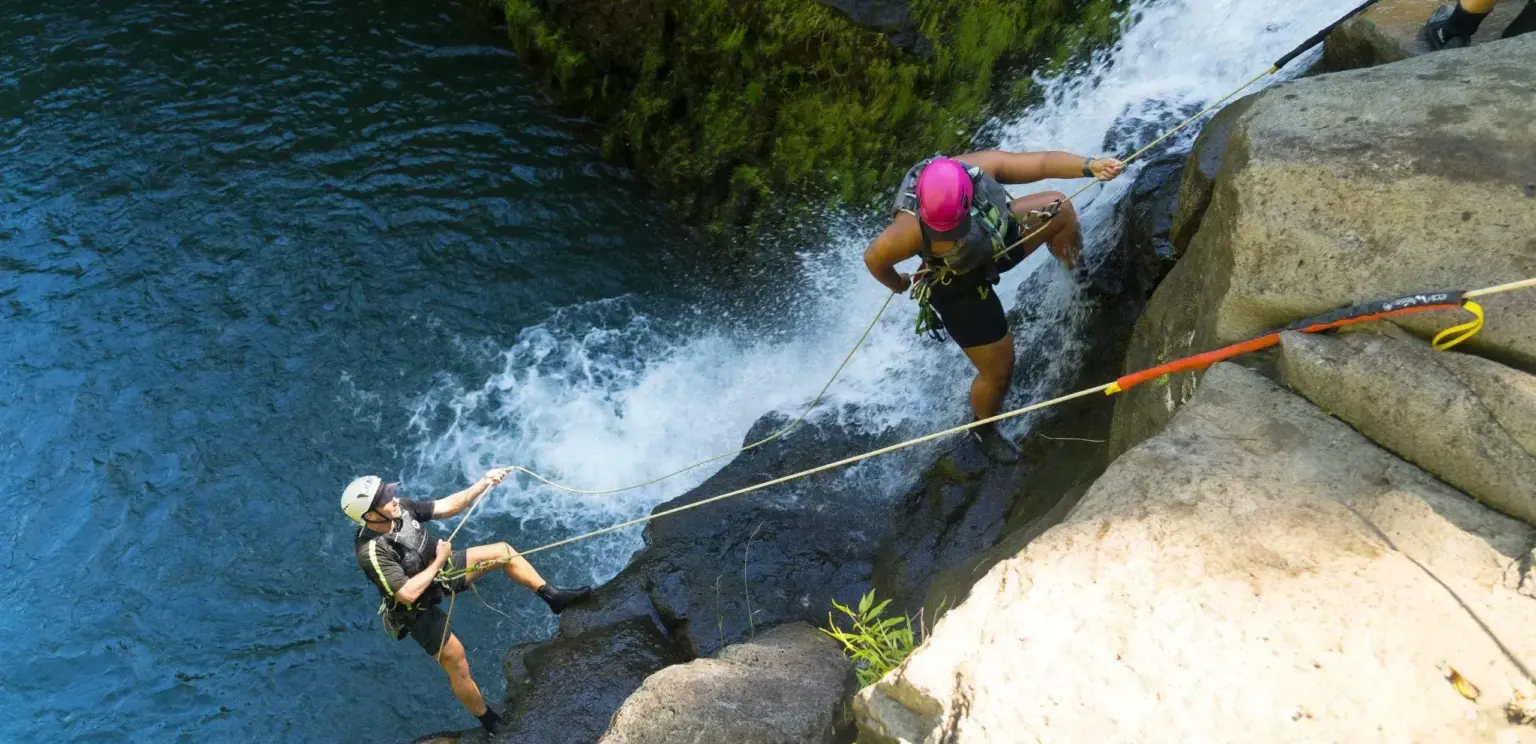 two people rappelling down a waterfall near mossy rocks over blue water