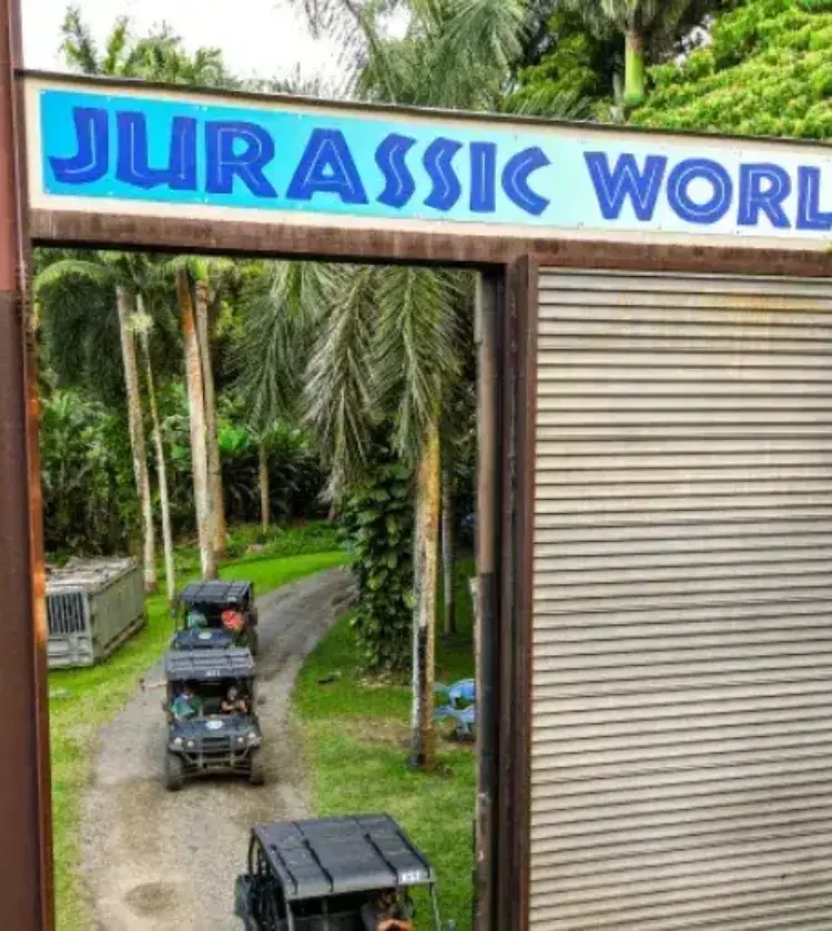 Jurassic World sign over entrance with safari vehicles on dirt road through tropical trees