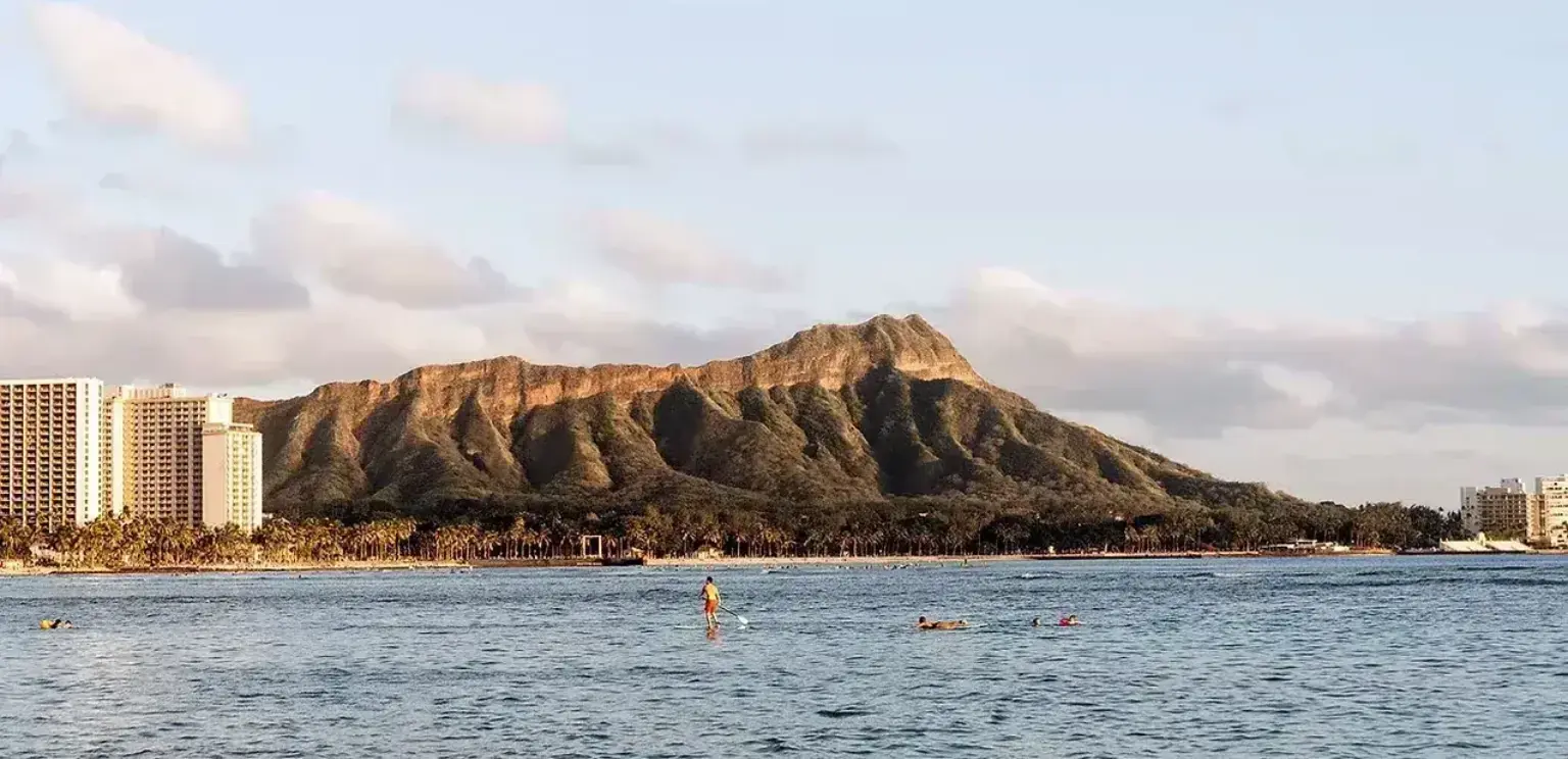 Diamond Head volcano in Honolulu with ocean and paddleboarders in foreground under cloudy sky