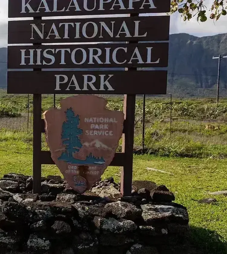 Kalaupapa National Historical Park wooden sign on stone base with green field and mountain range in the background