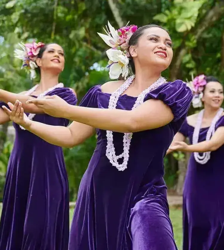Hawaiian hula dancers wearing purple dresses, floral headpieces, and white leis performing outdoors with green trees in background